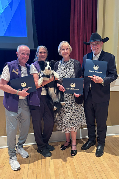 Navigator Jan Dougherty, Navigator Chaplain John Barelli, and Navigator Buddy team Beth and Will Thompson, along with their therapy dog Finn standing and holding their Impact Awards