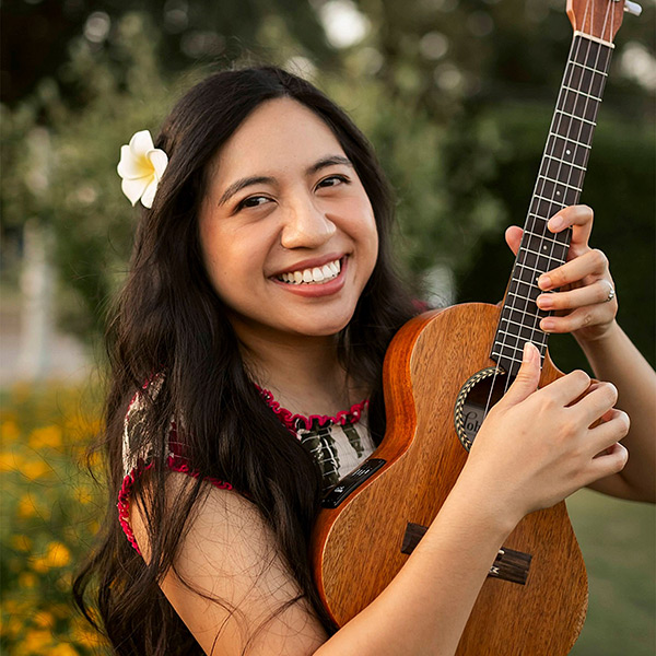 Jessica Maeve smiling at the camera with a flower in her hair while playing a ukulele
