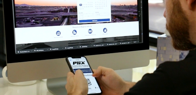 Man holding a cell phone in front of a computer monitor.