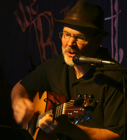 Performing artist Jim Sills playing the guitar and singing on a stage with dramatic lighting